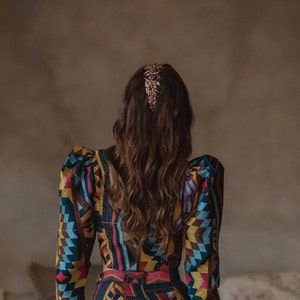 May include: A woman with long brown hair wearing a colorful patterned dress with puffed sleeves. She is wearing a gold hair comb.