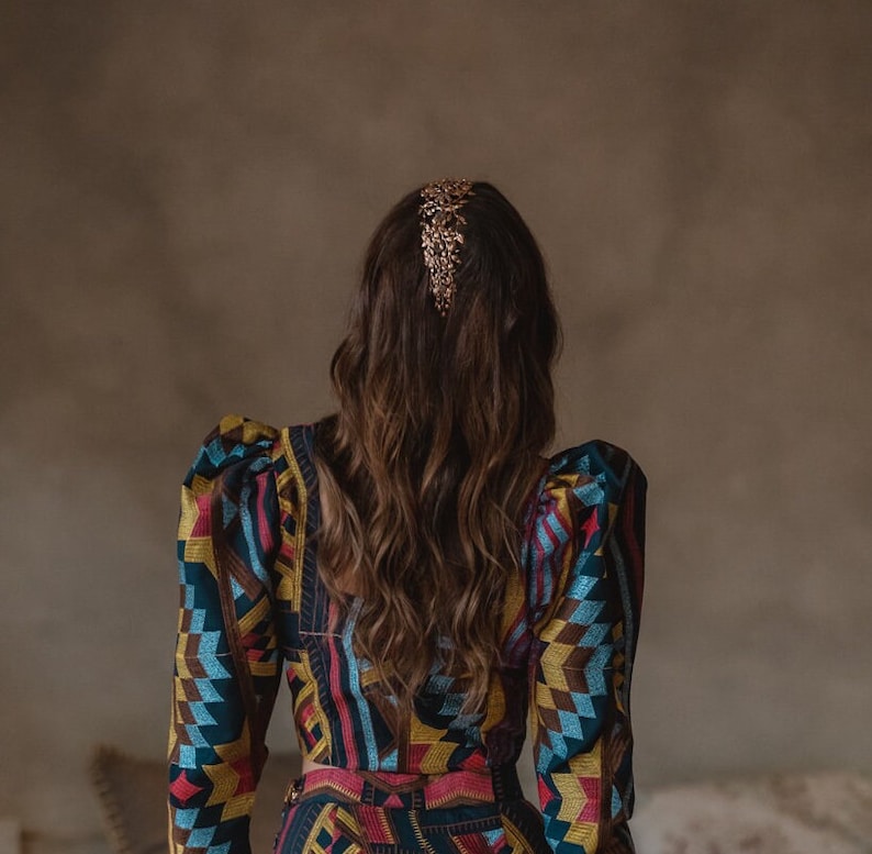 May include: A woman with long brown hair wearing a colorful patterned dress with puffed sleeves. She is wearing a gold hair comb.