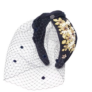 May include: A navy blue fabric headband featuring a knotted top and a sheer navy blue birdcage veil with small dark pom-poms. The headband is decorated with a cluster of metallic gold-toned floral embellishments and small white beads.