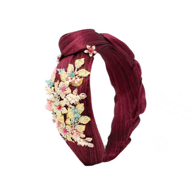 May include: A deep burgundy knotted headband featuring a textured fabric finish. The side is adorned with a cluster of metallic gold leaves, small pale pink flowers, and tiny light blue accents, creating a detailed floral arrangement on the band.