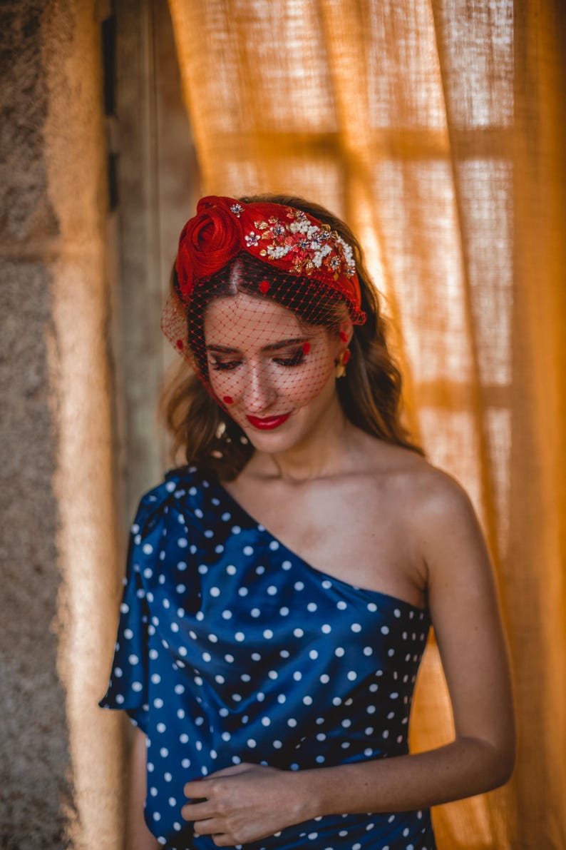 Puede incluir: Una diadema roja con una gran rosa de tela, intrincados abalorios florales y un velo negro tipo jaula con pequeños lunares rojos. La diadema se combina con un top azul marino de un solo hombro con un estampado de lunares blancos.
