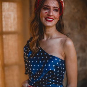 May include: A navy blue one-shoulder dress with white polka dots, paired with a red headband featuring a fine mesh veil and floral embellishments. A red rectangular clutch with a gold clasp and red feather accents is held in the front.
