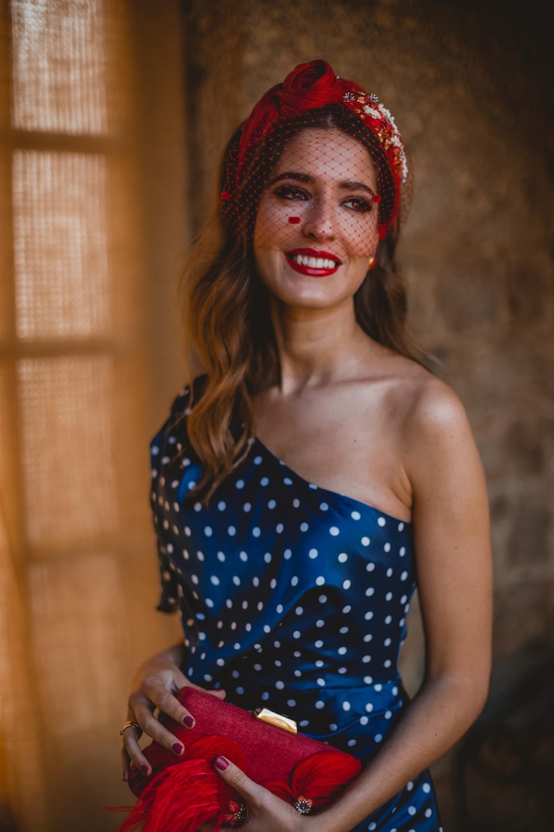 May include: A navy blue one-shoulder dress with white polka dots, paired with a red headband featuring a fine mesh veil and floral embellishments. A red rectangular clutch with a gold clasp and red feather accents is held in the front.