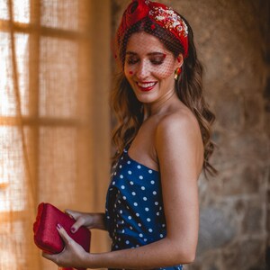 May include: A red headband adorned with floral embellishments and a black dotted veil, paired with a navy blue one-shoulder dress featuring white polka dots and a rectangular red clutch bag with a red feather tassel accent.
