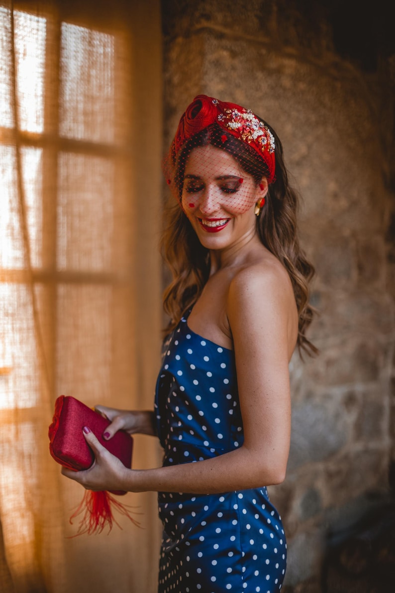 May include: A red headband adorned with floral embellishments and a black dotted veil, paired with a navy blue one-shoulder dress featuring white polka dots and a rectangular red clutch bag with a red feather tassel accent.