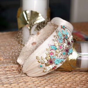 May include: A beige padded headband featuring a delicate beige netting veil. The side is adorned with an intricate floral arrangement of metallic gold leaves, turquoise blue petals, small white flowers, and vibrant pink accents.