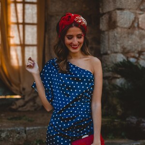 May include: A navy blue one-shoulder dress with a white polka dot pattern, paired with a red headband featuring floral embellishments and a fine mesh veil. A rectangular red clutch bag with a decorative red feather tassel is held in the hand.
