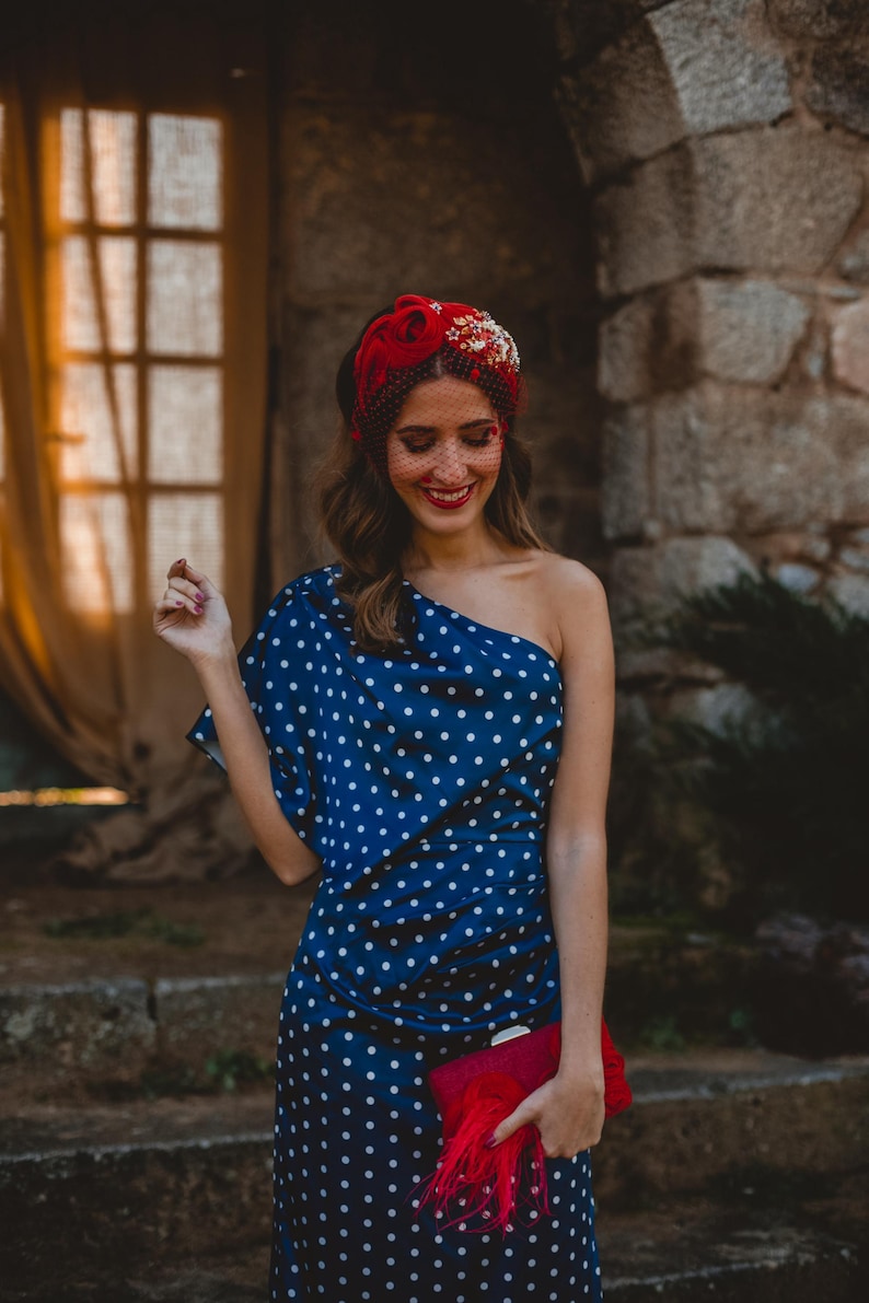 May include: A navy blue one-shoulder dress with a white polka dot pattern, paired with a red headband featuring floral embellishments and a fine mesh veil. A rectangular red clutch bag with a decorative red feather tassel is held in the hand.