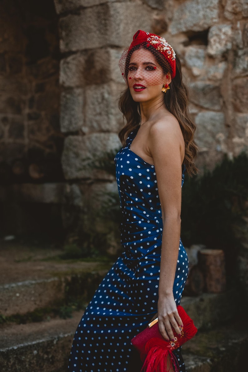 May include: A navy blue one-shoulder dress with a white polka dot pattern, paired with a red headband featuring floral embellishments and a red netting veil. A red clutch with decorative floral accents is held in the lower portion of the frame.