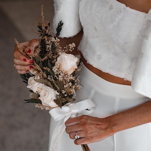 May include: A bride holds a bouquet of white roses and dried flowers with a white ribbon tied around the stems. She is wearing a white crop top and skirt.