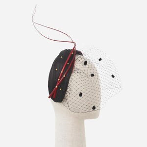 Könnte beinhalten: Schwarzes Kopfstück mit rotem Akzent und gepunktetem Schleier. Das Kopfstück ist mit kleinen goldfarbenen Verzierungen versehen. Ein dünnes rotes Band erstreckt sich von oben. Der Schleier ist schwarz mit schwarzen Punkten.