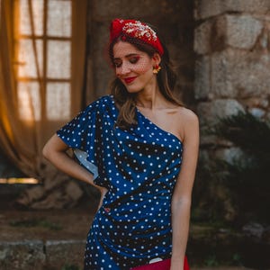 May include: A navy blue one-shoulder dress with a white polka dot pattern, paired with a red headband featuring floral embellishments and a red veil. A red clutch with feather accents is held in front of the dress, and gold flower earrings are worn.
