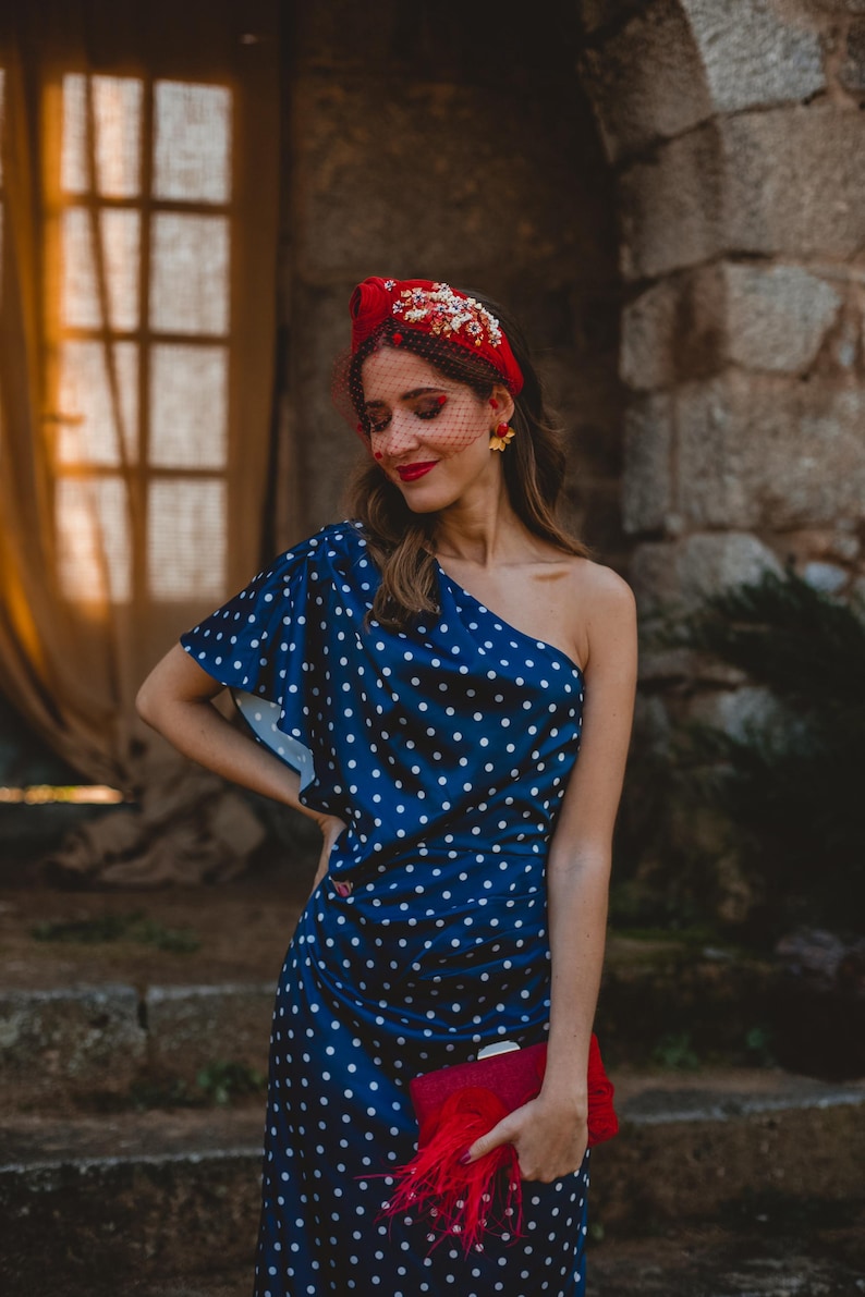 May include: A navy blue one-shoulder dress with a white polka dot pattern, paired with a red headband featuring floral embellishments and a red veil. A red clutch with feather accents is held in front of the dress, and gold flower earrings are worn.