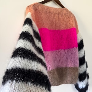 Puede incluir: Un suéter de gran tamaño y cuello redondo. El cuerpo del suéter presenta rayas horizontales en melocotón, rosa intenso, rosa claro y marrón. Las mangas son de rayas blancas y negras. El suéter está colgado en una percha de madera.