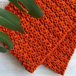 Może przedstawiać: Zbliżenie dwóch pomarańczowych kwadratów szydełkowych. Szydełko ma teksturowany wygląd, z widocznymi ściegami tworzącymi gęsty wzór. Kolor jest żywy, ciepły pomarańczowy. Kwadraty są umieszczone na białej drewnianej powierzchni, z zielonymi liśćmi w lewym górnym rogu.