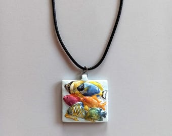 Può includere: Collana con pendente quadrato raffigurante un gruppo di pesci colorati. Il pendente ha uno sfondo bianco e un cordino nero. I pesci sono gialli, blu, arancioni, rosa e verdi.