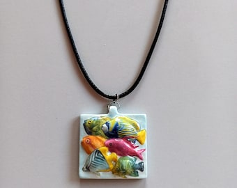 Può includere: Una collana con pendente quadrato con un rilievo di sei pesci tropicali colorati. I pesci sono in tonalità di giallo, arancione, rosa, verde e blu. Il pendente è attaccato a una collana con cordino nero.