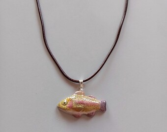 Può includere: Collana con pendente a forma di pesce. Il pendente è una rappresentazione dettagliata e colorata di un pesce, con sfumature gialle, rosa e viola. Pende da una collana di corda marrone con chiusura argentata. Lo sfondo è neutro.
