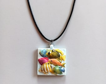Può includere: Collana con pendente quadrato raffigurante pesci tropicali colorati. Il pendente, con sfondo bianco e anello argentato, è sospeso a una collana di corda nera. I pesci sono dipinti in giallo, arancione, rosa e blu.