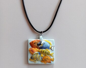 Può includere: Collana con pendente quadrato azzurro con pesci colorati. I pesci sono arancioni, gialli e blu. La collana ha un cordino nero.