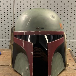 May include: A weathered green and maroon helmet with a black visor, reminiscent of a bounty hunter's headgear. The helmet features orange stripes and a rangefinder. The design suggests a science fiction or cosplay theme.