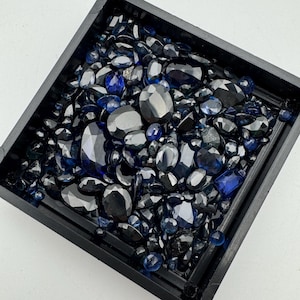 May include: A square black tray filled with an assortment of dark blue and black gemstones. The gemstones vary in size and shape, including oval, round, and faceted cuts. The gems are arranged closely together, creating a dense, textured surface.