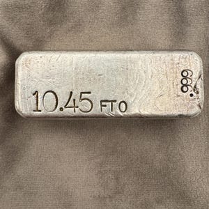 May include: A silver bar with the stamped text "10.45 FTO" and "999" on the surface. The bar has a rectangular shape with rounded edges. The background is a neutral, textured fabric.