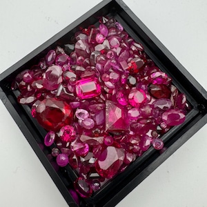 May include: A square black tray filled with a variety of red and pink gemstones. The gemstones are of different shapes and sizes, including round, oval, and rectangular cuts. The gems vary in shades from deep red to light pink.