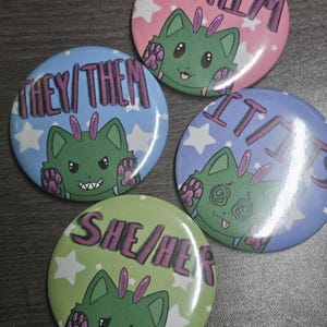 May include: Four circular pin-back buttons featuring green cat-like creatures with purple accents. Each button displays different pronouns in black text: HE/HIM on pink, THEY/THEM on blue, IT/ITS on purple, and SHE/HER on light green, all with white stars.