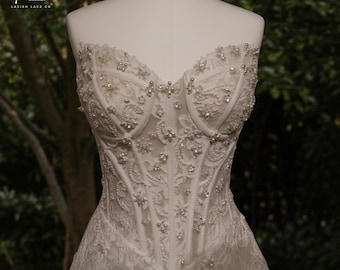 May include: A white, strapless corset-style bodice with intricate lace detailing and beaded embellishments. The bodice is displayed on a mannequin, showcasing the delicate floral patterns and structured design. The image is set against a blurred green background.