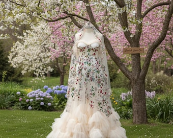 May include: A white wedding dress with floral embroidery, displayed on a mannequin outdoors. The dress features a sweetheart neckline, a fitted bodice, and a layered tulle skirt. A sheer veil is draped over the mannequin's head. The background includes trees with pink and white blossoms.