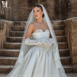 Puede incluir: Un vestido de novia blanco sin tirantes con falda amplia y detalles de encaje. La novia lleva guantes largos de encaje y un velo transparente. El texto "LAVISH LACE CO" y "REAL IMAGES BY LAVISH COUTURE" está presente.