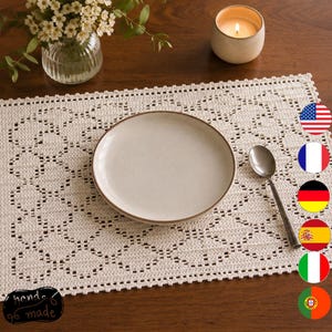 May include: A rectangular off-white crocheted placemat featuring an intricate openwork geometric pattern. The placemat is centered on a wooden surface with a plain ceramic plate and a silver spoon placed on top of the textile surface.