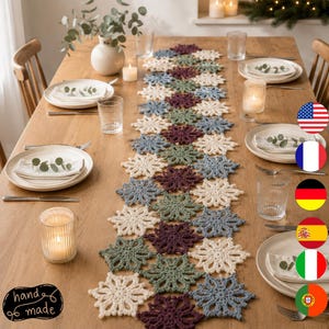 Puede incluir: Un camino de mesa largo compuesto por motivos de copos de nieve tejidos a ganchillo en tonos crema, verde salvia, azul polvoriento y ciruela oscuro. El camino está centrado sobre una mesa de madera con platos blancos, servilletas y velas.