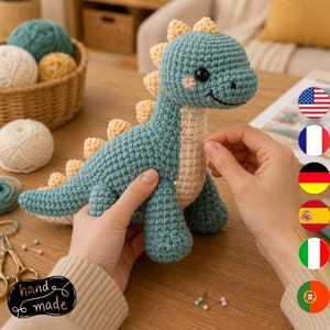 Puede incluir: Un juguete de dinosaurio tejido a ganchillo en color verde azulado y crema con una hilera de pequeñas púas amarillas en la espalda. El dinosaurio tiene ojos de seguridad negros, una sonrisa bordada y detalles rosados en las mejillas.