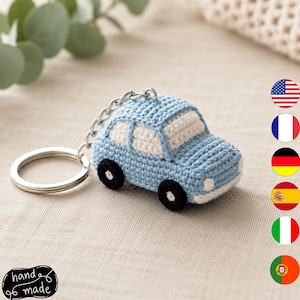 Puede incluir: Un llavero de coche tejido a ganchillo en color azul claro con ventanas blancas, ruedas negras y una anilla de metal plateado. El coche está elaborado con un punto de hilo texturizado, mostrando un diseño compacto con parachoques y faros.