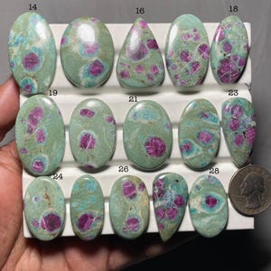 May include: A selection of polished gemstones in diverse shapes, such as oval and teardrop. The stones display a mottled pattern of green, teal, and deep pink or red hues. A coin is included for size reference. The gemstones are presented on a white surface.