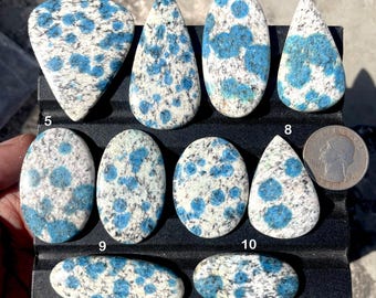 May include: A collection of K2 stone cabochons in various shapes, including teardrop and oval. The stones feature a white base with striking blue orb-like patterns. A quarter is included for size reference. These polished stones are ideal for jewelry making.