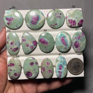 Puede incluir: Una colección de piedras preciosas pulidas de varias formas, incluyendo ovaladas y redondas. Las piedras presentan un color base verde moteado con inclusiones magenta vibrantes. Se incluye una moneda para referencia de tamaño.