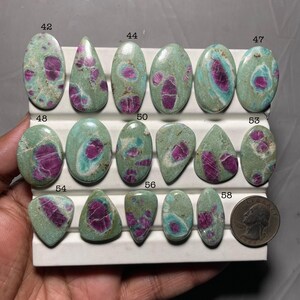 Puede incluir: Una colección de piedras preciosas pulidas con una combinación de colores turquesa y magenta. Las piedras tienen varias formas, incluyendo ovaladas, de lágrima y triangulares. Se incluye una moneda para la comparación de tamaño. Las piedras están numeradas.