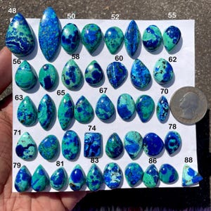 May include: A selection of polished azurite gemstones in diverse shapes, such as teardrops and ovals. The stones exhibit a blend of deep blue and teal green colours, with a quarter coin for size reference. The gemstones are numbered.