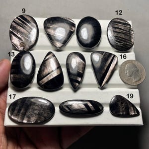 May include: Assortment of polished black gemstones with iridescent silver inclusions, displayed on a white tray. Various shapes include ovals, teardrops, and triangles. A quarter is included for size reference. The gemstones are likely for jewelry making.