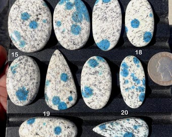 May include: A collection of polished k2 azurite stones in various oval and teardrop shapes. The stones feature a white and gray granite base with vibrant blue azurite inclusions. A US quarter is included for size reference.