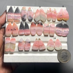 Puede incluir: Una colección de cabujones de piedras preciosas rosas y grises en varias formas, incluyendo óvalos, lágrimas y diseños puntiagudos. Las piedras se exhiben sobre una superficie blanca, con una moneda de veinticinco centavos para referencia de tamaño. Las piedras tienen patrones únicos.