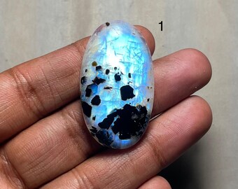 May include: An oval-shaped moonstone gemstone with a pearlescent sheen. The stone features a mix of blue, white, and black inclusions. The gemstone is held in a hand, showcasing its size and color variations.