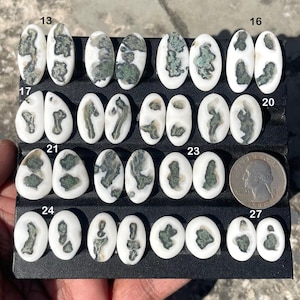 May include: A collection of oval-shaped gemstones with white and green patterns. The gemstones are arranged on a black surface, with a US quarter for size comparison. The stones vary in size and pattern.