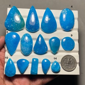 May include: A collection of polished, vibrant blue gemstones in various shapes, including teardrops, ovals, and triangles. The gemstones are displayed on a white tray, with a quarter coin for size comparison. The stones have a smooth, glossy finish.