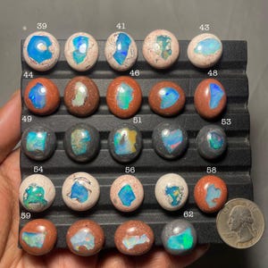 May include: A collection of polished opal cabochons in various colors, including blue, green, and orange, set against a black background. Each stone is numbered, showcasing the unique patterns and hues of the opals. A quarter is included for size reference.