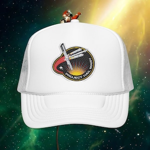 May include: White trucker hat with a mesh back and a curved brim. The front features an embroidered patch with the text "PROJECT HAIL MARY" and a graphic of a spaceship and sunburst. A small astronaut figure sits on top.