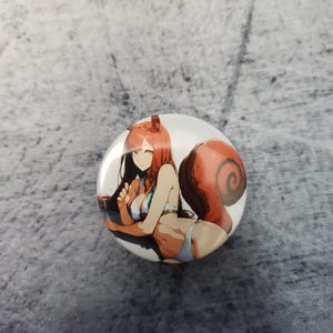 May include: Round button with a white background featuring an anime-style illustration of a woman with red hair and a large, fluffy, red squirrel tail. She is wearing a blue bikini. The button is on a gray surface.